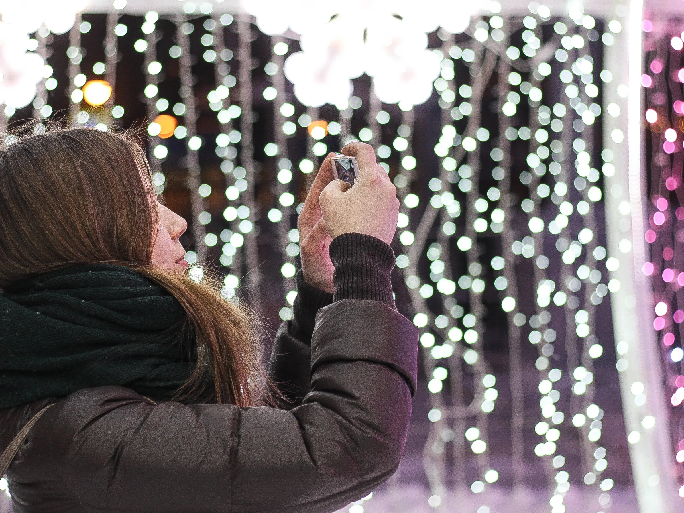 A woman filming with her phone in front of a wall of string lights